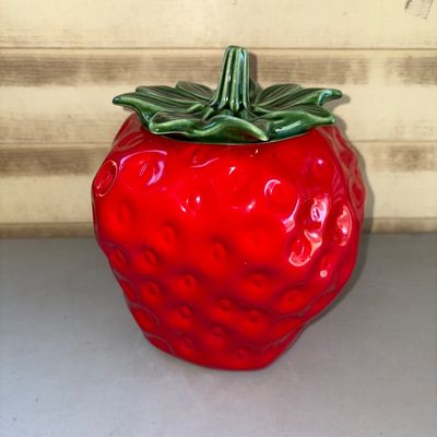 Sale Photo Thumbnail #172: Ceramic strawberry-shaped cookie jar featuring a bright red glazed body with textured seed dimples and a green leaf-and-stem lid. Features a glossy glazed interior. Good pre-owned condition with no visible cracks or large chips.
11in tall