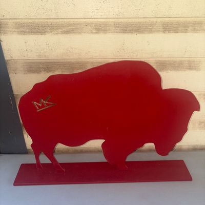 Sale Photo Thumbnail #249: A painted red metal buffalo or bison tabletop silhouette mounted on a flat base. Features a gold-toned logo/brand emblem near the top left hip. Overall good condition with minor surface wear.
13.5X20X3.5in