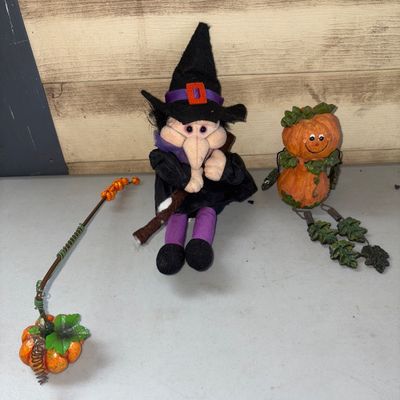 Sale Photo Thumbnail #366: A set of 3 Halloween decor items featuring a plush witch figurine holding a broom, a shelf-sitter pumpkin figure with leaf-style dangling legs, and a small decorative pumpkin stake accent with wire and beads. Items show minor wear, dust, and lint consiste
