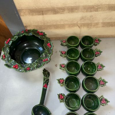 Sale Photo Thumbnail #338: A ceramic holiday punch bowl set featuring a green glazed finish with raised holly leaf and red berry accents around the rims and handles. Includes 1 large punch bowl, 1 ceramic ladle, and 12 matching cups. Minor wear visible, including a small chip on on