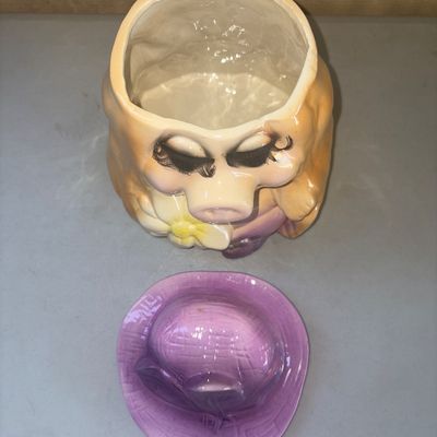 Sale Photo Thumbnail #248: Vintage ceramic cookie jar featuring Miss Piggy wearing a purple hat and holding a white and yellow daisy. Includes removable purple hat lid. Overall good condition with glossy finish; minor wear and light paint imperfections consistent with age and use.

