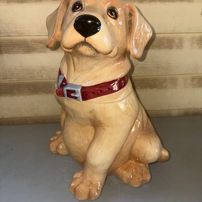 Sale Photo Thumbnail #256: Glazed ceramic cookie jar shaped like a sitting yellow Labrador Retriever wearing a red collar. Features a removable head lid. Shows light crazing in the glaze visible on the body; otherwise in good condition with no apparent chips or cracks.
12in tall