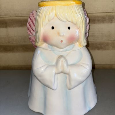 Sale Photo Thumbnail #164: Charming ceramic praying angel cookie jar with lid. Features pastel blue and white robes, blonde hair with a yellow halo, and pink feathered wings. Glazed finish. Shows light wear consistent with age, including subtle hairline cracking near the top rim of