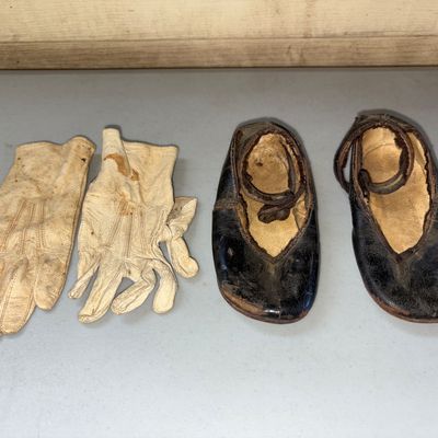 Sale Photo Thumbnail #6: A pair of antique leather child or doll shoes with ankle straps alongside a pair of stitched cream-colored leather gloves. Items show significant age-related wear, patina, surface cracking, and staining consistent with vintage/antique age.