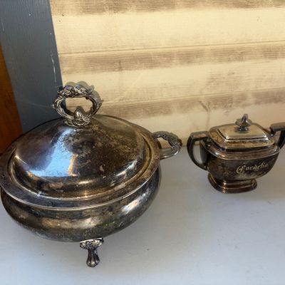 Sale Photo Thumbnail #265: A 2-piece collection of silver-plated serveware including a large footed covered tureen with ornate scroll handles and top finial, and a small dual-handled covered sugar bowl engraved with 'Cordelia'. Both pieces feature significant heavy tarnish, patina,