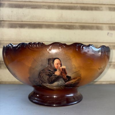 Sale Photo Thumbnail #383: Large antique ceramic or earthenware footed bowl featuring a scalloped rim and a shaded brown-to-tan glaze. Decorated with hand-painted or transfer floral poppy designs on one side and an interior, and a portrait of a monk drinking beer/ale on the opposit