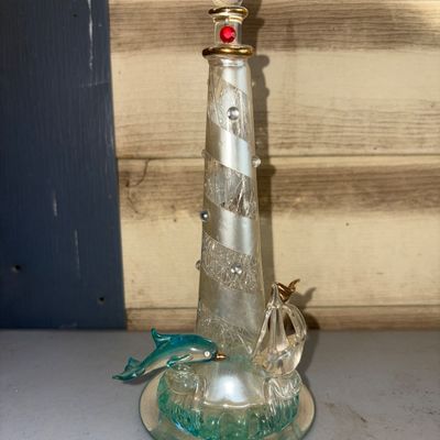 Sale Photo Thumbnail #348: Decorative blown glass lighthouse sculpture featuring crackle glass texture with frosted spiral stripes, clear applied cabochons, gold-tone accents, and a red rhinestone jewel near the top. The base includes a teal-accented wave motif with a leaping teal 