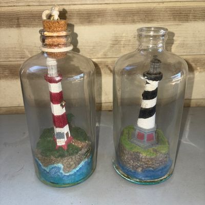 Sale Photo Thumbnail #146: Pair of detailed nautical miniature lighthouse figures enclosed in glass bottles. Includes one red-and-white striped lighthouse with cork stopper and rope detail, and one black-and-white spiral lighthouse. Note: The base of the black and white lighthouse 