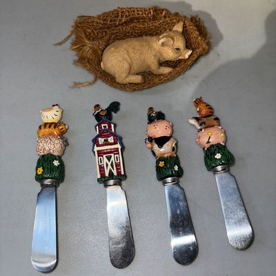 Sale Photo Thumbnail #60: A country-themed set featuring four stainless steel cheese spreaders with resin novelty handles (stacked barnyard animals and red barn motif) alongside a small decorative pig figurine resting in a textured burlap sack. All pieces are in good pre-owned con