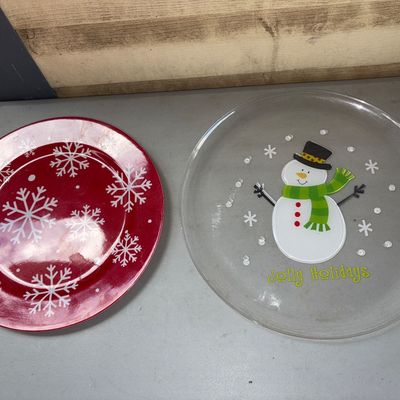 Sale Photo Thumbnail #318: Includes two round plastic Christmas holiday serving plates. One features a clear glass construction with a central snowman graphic and 'Jolly Holidays' text with snowflake accents. The second plate features a red finish with white snowflake patterns thro