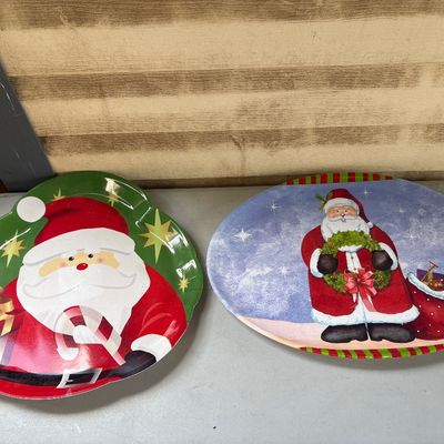 Sale Photo Thumbnail #319: Set of two festive plastic/melamine Christmas serving trays featuring Santa Claus designs. One oval tray with a blue snowy background shows noticeable damage and chipping along the right edge. The second green scalloped tray features Santa holding a prese