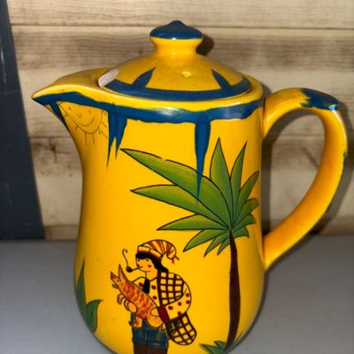 Sale Photo Thumbnail #217: Vibrant yellow ceramic pitcher or teapot featuring colorful hand-painted folk art illustrations including a figure with a pipe under a palm tree and a smiling sun motif. Bright blue glaze accents trim the spout, rim, lid, and handle. Note: There is a nota