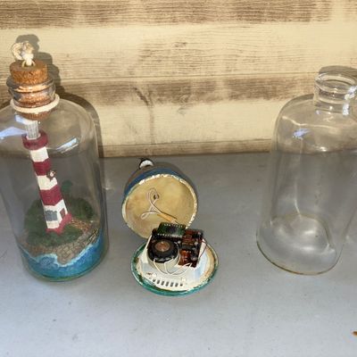 Sale Photo Thumbnail #147: Pair of detailed nautical miniature lighthouse figures enclosed in glass bottles. Includes one red-and-white striped lighthouse with cork stopper and rope detail, and one black-and-white spiral lighthouse. Note: The base of the black and white lighthouse 