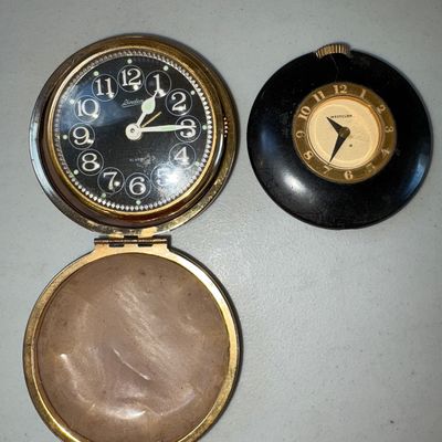 Sale Photo Thumbnail #7: Pair of vintage compact clocks including a Linden folding travel alarm clock in a gold-tone and black clam-shell compact case with a fabric lining, and a small round Westclox pocket/desk clock in a black case. Items show age-appropriate surface wear, scuf
