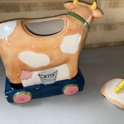 Sale Photo Thumbnail #88: Charming hand-painted ceramic cookie jar featuring a cow on a pull-toy style base with a cat, milk pail detail, and a crescent moon lid handle inspired by the nursery rhyme. Shows minor paint wear and small chips on the moon handle tip and horn.
10X8X10in
