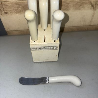 Sale Photo Thumbnail #294: Set of off-white handled cheese spreaders in a matching storage block with floral border detail. Set includes 5 spreaders in the block with one shown removed (total of 6 slots in block, 5 present). Shows some light discoloration, scuffing, and minor wear 