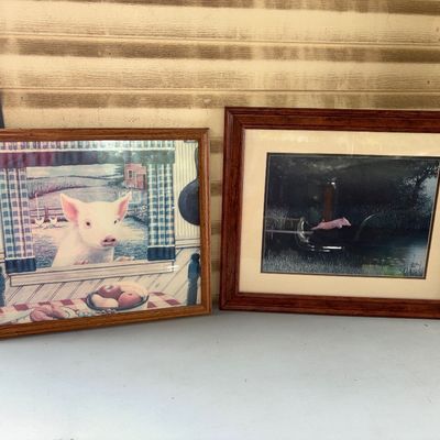 Sale Photo Thumbnail #106: Set of two framed pig-themed art prints. One features a pig looking through a kitchen window in a country setting, and the other shows a flying pig over a nocturnal lake scene. Both come encased in wooden frames behind glass or plastic face protection. In