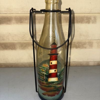 Sale Photo Thumbnail #203: Decorative amber glass bottle featuring a hand-painted style red-and-white striped lighthouse scene with birds and coastal landscape. Held within a black wrought-iron style wire caddy stand with handle. Appears to be in good condition with minor surface w