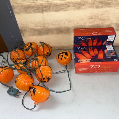 Sale Photo Thumbnail #375: Assortment of Halloween lights including a strand of jack-o'-lantern pumpkin blow mold style string lights and two boxed Hobby Lobby 70-count super bright orange light sets. Items appear to be in good used condition. Boxes show light shelf wear.