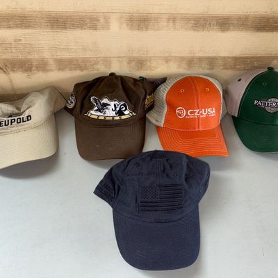 Sale Photo Thumbnail #96: Collection of 5 adult baseball caps and hats featuring outdoor, hunting, and firearms branding including Leupold (khaki visor/cap), CZ-USA (orange mesh-back trucker cap), Patternmaster (green/tan), a brown embroidered wildlife cap dated 2005, and a navy b