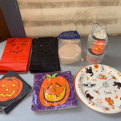Sale Photo Thumbnail #363: An assortment of Halloween party table goods and decorations. Includes cut-out jack-o'-lantern paper bags (orange and black), paper napkins featuring pumpkin designs, white crepe paper streamer rolls, a stack of paper cups, and a decorative Halloween pape