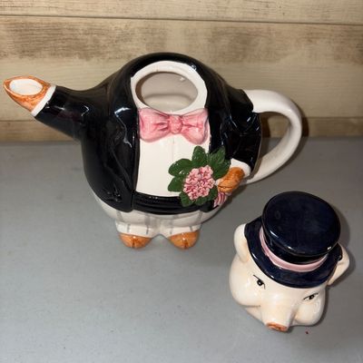 Sale Photo Thumbnail #55: Novelty ceramic teapot shaped like a dapper pig dressed in a black top hat, tuxedo coat, pink bow tie, and holding a small bouquet of flowers. Features a spout formed by one arm and a rear handle. Appears to be in good pre-owned condition with no visible 