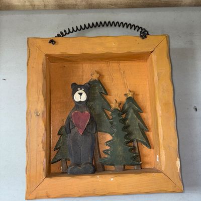 Sale Photo Thumbnail #83: 8X8.5in
Folk art style wooden shadow box featuring a cutout bear holding a red heart standing among evergreen trees topped with stars. Housed in a yellow-painted rustic wood frame with a coiled wire hanging hanger at the top. Shows light wear and minor sc