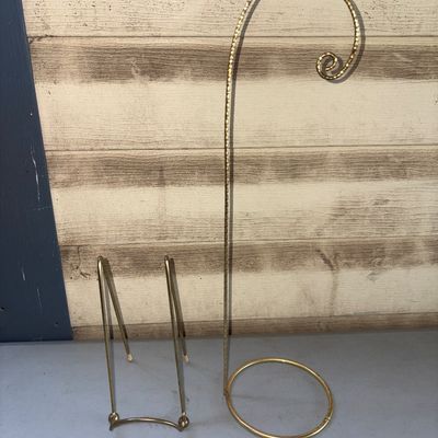 Sale Photo Thumbnail #298: Pair of brass-toned metal display stands. Includes one tall hanging ornament or pocket watch display hanger with a twisted metal stem and round base, and one folding wire plate or book easel stand. Both items show light wear consistent with gentle use.
6i
