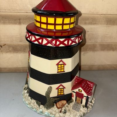 Sale Photo Thumbnail #89: Hand-painted ceramic lighthouse cookie jar or decorative storage canister. Features a black and white striped body with a red roof, yellow lit-window accents, and a rocky shoreline base with a small attached keeper's house. In good condition with vibrant 