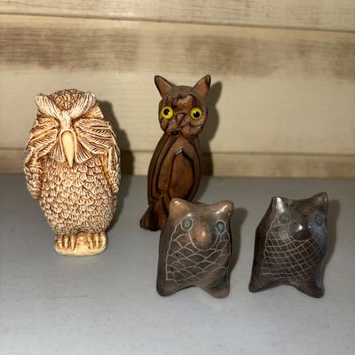Sale Photo Thumbnail #17: A collection of four owl figurines crafted from various materials, including carved wood, molded resin/composite, and carved stone/soapstone. Set includes a detailed textured cream resin owl, a slender carved wooden owl with inset yellow glass eyes, and t