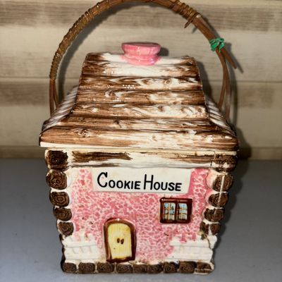 Sale Photo Thumbnail #180: Vintage ceramic cottage-style cookie jar labeled 'Cookie House'. Features a painted log cabin design with pink accents, a removable roof lid, and a woven wicker overhead handle. Shows some visible crazing, minor wear, and a hairline crack visible along th