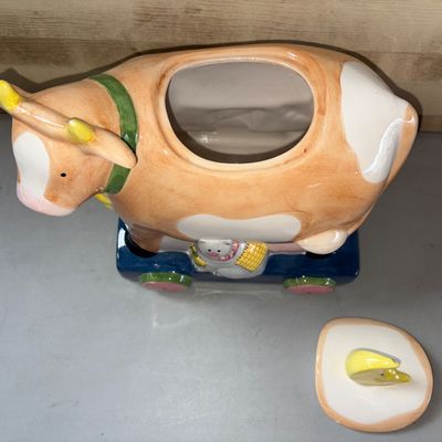 Sale Photo Thumbnail #85: Charming hand-painted ceramic cookie jar featuring a cow on a pull-toy style base with a cat, milk pail detail, and a crescent moon lid handle inspired by the nursery rhyme. Shows minor paint wear and small chips on the moon handle tip and horn.
10X8X10in