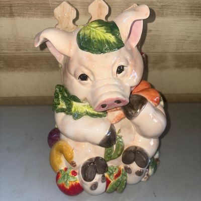 Sale Photo Thumbnail #258: Hand-painted ceramic cookie jar featuring a seated pig holding a carrot, surrounded by garden vegetables such as lettuce, tomatoes, and peas, with a picket fence detail on the back. The top section detaches as the lid. Glossy glazed finish. Shows light su