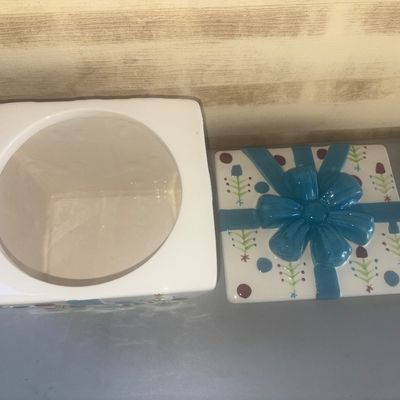 Sale Photo Thumbnail #238: Ceramic cookie jar or covered vessel shaped like a wrapped present, featuring a hand-painted style Christmas tree and polka dot motif on a white background, topped with a dimensional blue ceramic bow lid. Appears to be in good condition with no visible ch