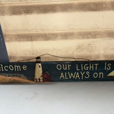Sale Photo Thumbnail #309: Decorative wooden hanging sign featuring a nautical theme with a lighthouse, sailboat, and the text 'Welcome - our light is always on'. Includes wire loop for easy hanging. Shows light wear consistent with display.
24X3.5in
