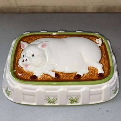 Sale Photo Thumbnail #92: Ceramic or stoneware mold featuring a raised relief pig design against a textured brown background with a white picket fence detail around the border. Includes a hanging hole on the back rim for display. Light wear consistent with age, glossy glaze finish