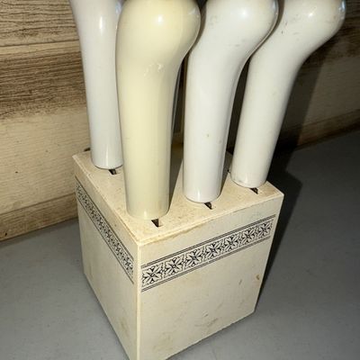 Sale Photo Thumbnail #293: Set of off-white handled cheese spreaders in a matching storage block with floral border detail. Set includes 5 spreaders in the block with one shown removed (total of 6 slots in block, 5 present). Shows some light discoloration, scuffing, and minor wear 