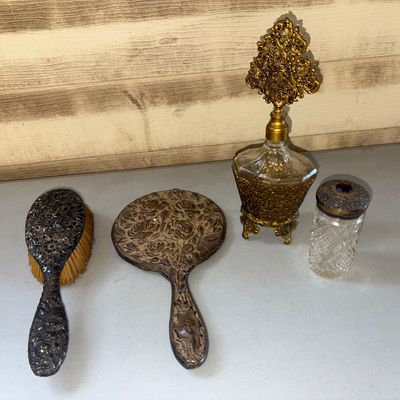 Sale Photo Thumbnail #9: A 4-piece vintage vanity collection featuring an ornate embossed metal hand mirror with a floral design on the reverse, a matching natural bristle hairbrush, an ornate brass or gold-tone filigree perfume bottle with a decorative stopper, and a cut glass v