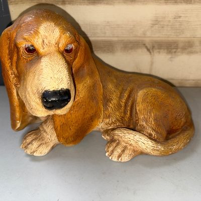 Sale Photo Thumbnail #322: A cast resin or composite sculpture depicting a resting Basset Hound with long floppy ears and textured fur details. Shows minor paint wear and light surface dust consistent with age. Measures 8in tall