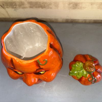 Sale Photo Thumbnail #156: Ceramic or pottery pumpkin cookie jar featuring a smiling jack-o'-lantern face and a removable lid styled with a stem and green leaf detail. Bright orange glazed finish with a white interior. Appears in good pre-owned condition with no visible chips or ma