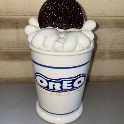 Sale Photo Thumbnail #195: A collectible ceramic Oreo cookie jar featuring a milk glass / milkshake design with whipped cream and a giant Oreo cookie on the lid. Front is decorated with the classic blue Oreo logo and the tagline "America's Favorite Cookie". The container is two pie