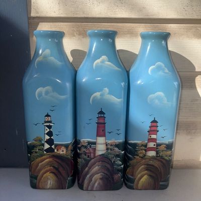 Sale Photo Thumbnail #343: A set of three light blue glass bottles or decorative vases, each featuring hand-painted nautical scenes with different lighthouses, rocky shores, clouds, and birds. Items appear to be in good pre-owned condition with no visible cracks or chips.
11in tall