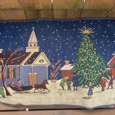 Sale Photo Thumbnail #316: A woven tapestry-style textile rug/mat featuring a festive winter village scene with a church, horse-drawn sleigh, decorated Christmas tree, and townspeople in the snow. Finished with a gray fabric border. Shows minor wear along the edges and light discol