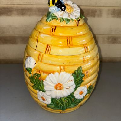 Sale Photo Thumbnail #174: Ceramic beehive-shaped cookie jar or canister featuring relief daisy flowers and a bumblebee finial on the lid. Bright yellow glazed finish with hand-painted details. Good condition with minor surface wear consistent with age and use.
10in tall