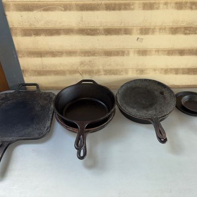 Sale Photo Thumbnail #395: Collection of various cast iron cookware including a square griddle, multiple round skillets of different sizes, and small miniature/ashtray skillets. Items show signs of prior use, surface residue, and oxidation typical of pre-owned cast iron, and may be