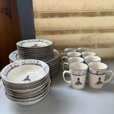 Sale Photo Thumbnail #306: A comprehensive ceramic dinnerware collection featuring a coastal lighthouse pattern with sailboat borders. The set includes large dinner plates, salad/salad plates, soup/cereal bowls, and matching coffee mugs. Items appear to be in good used condition wi