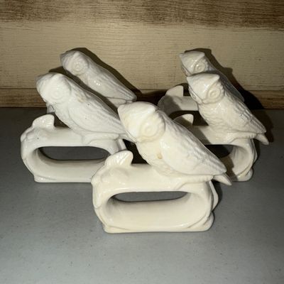 Sale Photo Thumbnail #77: A set of five white ceramic or porcelain napkin rings featuring sculpted owl figures perched on branches. Glossy finish. Overall good condition with light surface wear and minor discoloration consistent with age and use. Small visible cracks on one but in