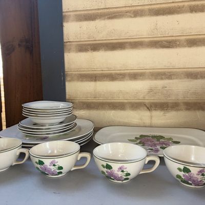 Sale Photo Thumbnail #303: A set of vintage cream-colored ceramic dinnerware featuring a hand-painted or printed purple floral violet pattern with green trim. The lot includes dinner plates, small bowls/saucers, a large serving platter, and 4 teacups. Items show typical vintage wea