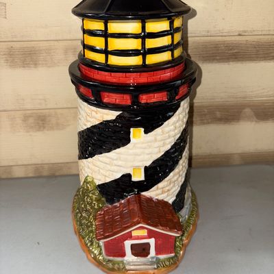 Sale Photo Thumbnail #90: A detailed ceramic cookie jar shaped like a classic black-and-white striped lighthouse featuring a red keeper's house at the base and a glazed top lid. The jar is in good condition with vibrant hand-painted details and no visible chips or cracks.
12X5in