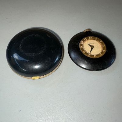 Sale Photo Thumbnail #8: Pair of vintage compact clocks including a Linden folding travel alarm clock in a gold-tone and black clam-shell compact case with a fabric lining, and a small round Westclox pocket/desk clock in a black case. Items show age-appropriate surface wear, scuf